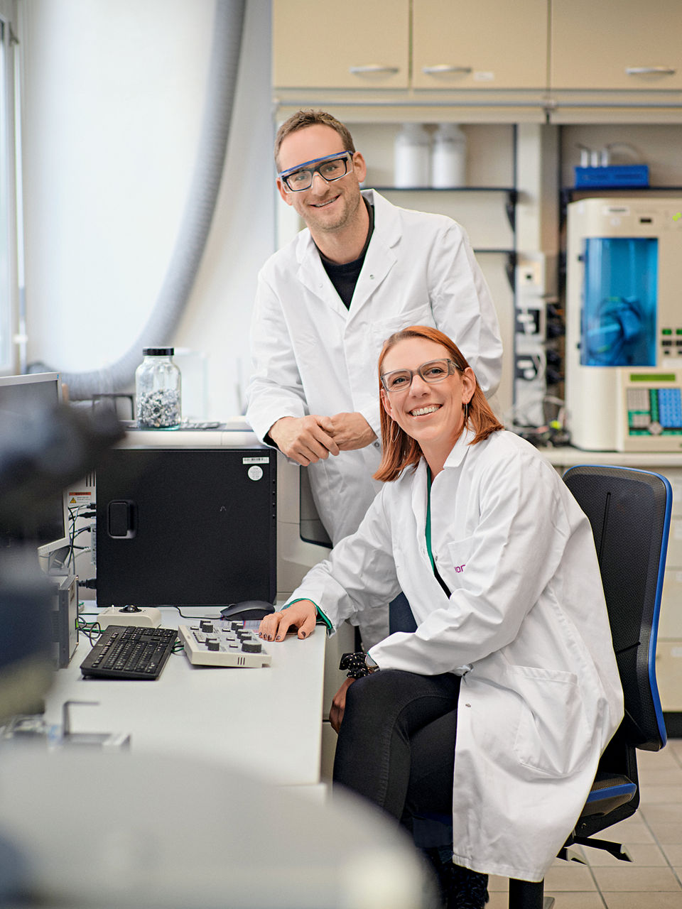 Max Braun und Jasmin Reiner gemeinsam an einem Schreibtisch. Beide tragen Laborkittel.