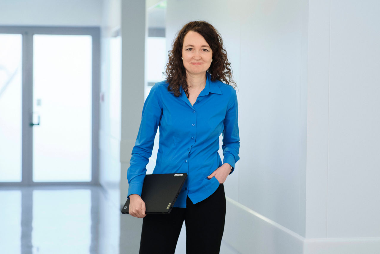 Bettina Hölzer steht mit einem Laptop in der Hand in einem hellen Flur und schaut in die Kamera.