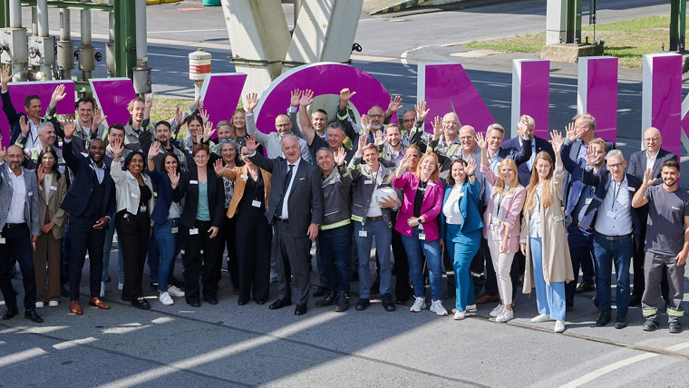 Large group of diverse professionals waving, standing in front of big purple letters spelling a word outdoors.