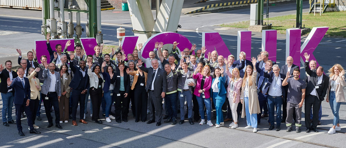 Large group of diverse professionals waving, standing in front of big purple letters spelling a word outdoors.