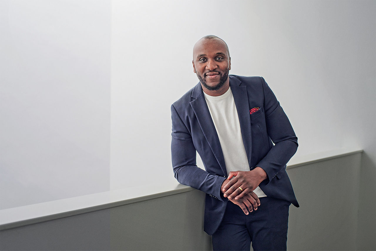 Christian Mani lehnt leicht seitlich an einem Geländer. Er trägt einen blauen Anzug und ein weißes T-Shirt.