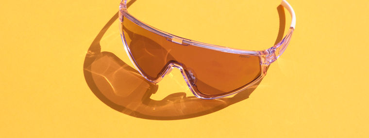 Protective eyewear with amber-tinted lenses and a transparent frame placed on a vibrant yellow background. The safety glasses feature a wraparound design for full eye coverage, ideal for industrial, laboratory, or outdoor use.