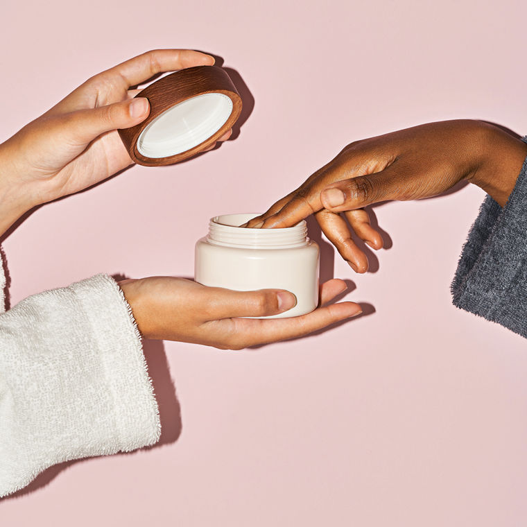 Zwei Frauenhände öffnen von links einen Creme-Tiegel. Eine Frauenhand nimmt von rechts mit einem Finger etwas aus dem Tiegel.