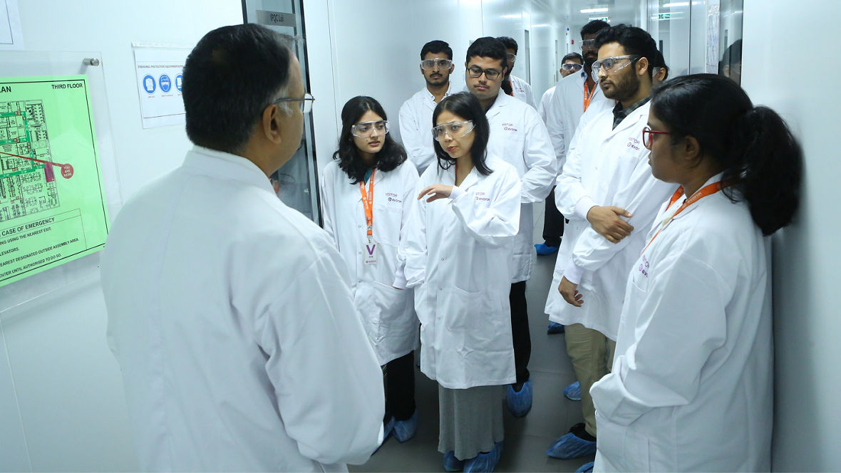 A group of individuals in lab coats attends a presentation in a corridor, engaged with a speaker.