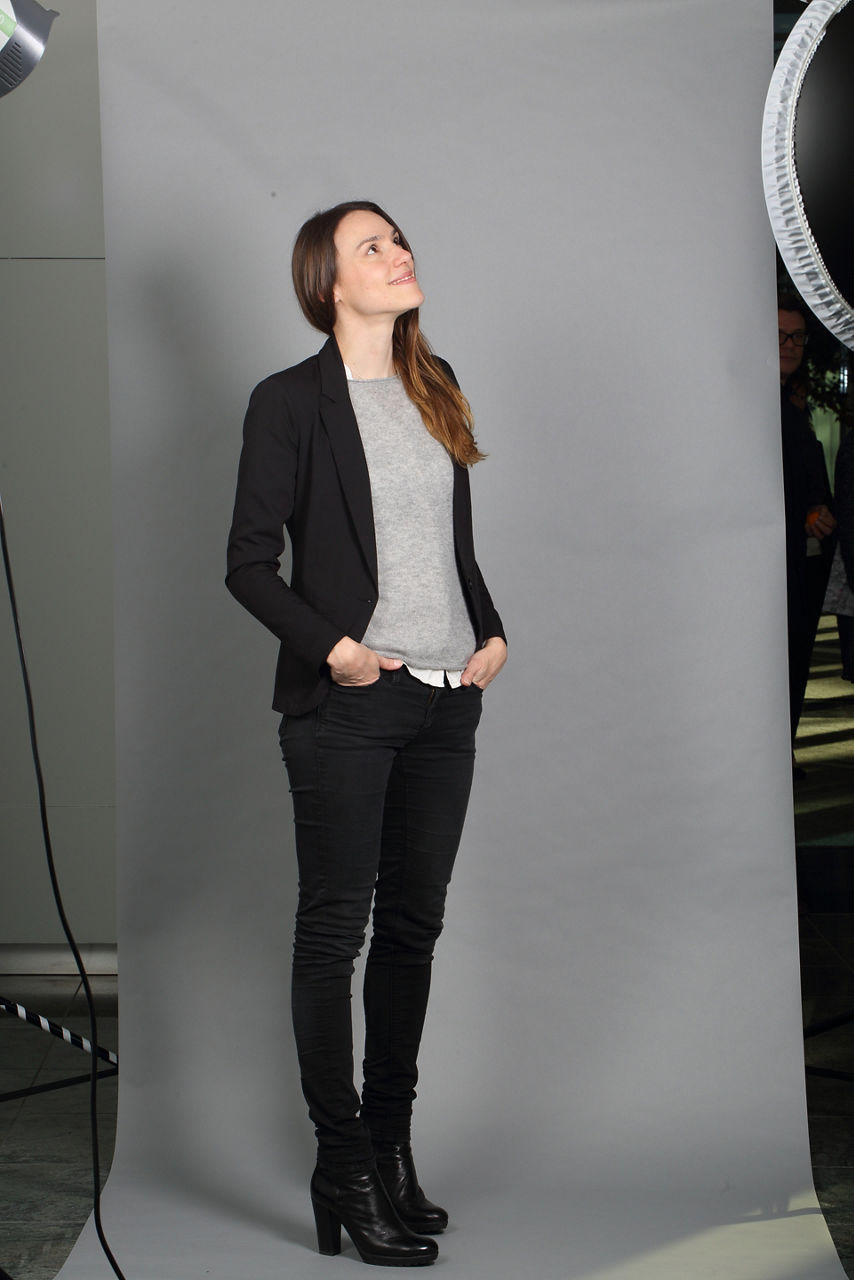 Sarah Hintermayer steht, mit den Händen in den Hosentaschen, vor einem grauen Hintergrund und schaut lächelnd nach rechts oben.