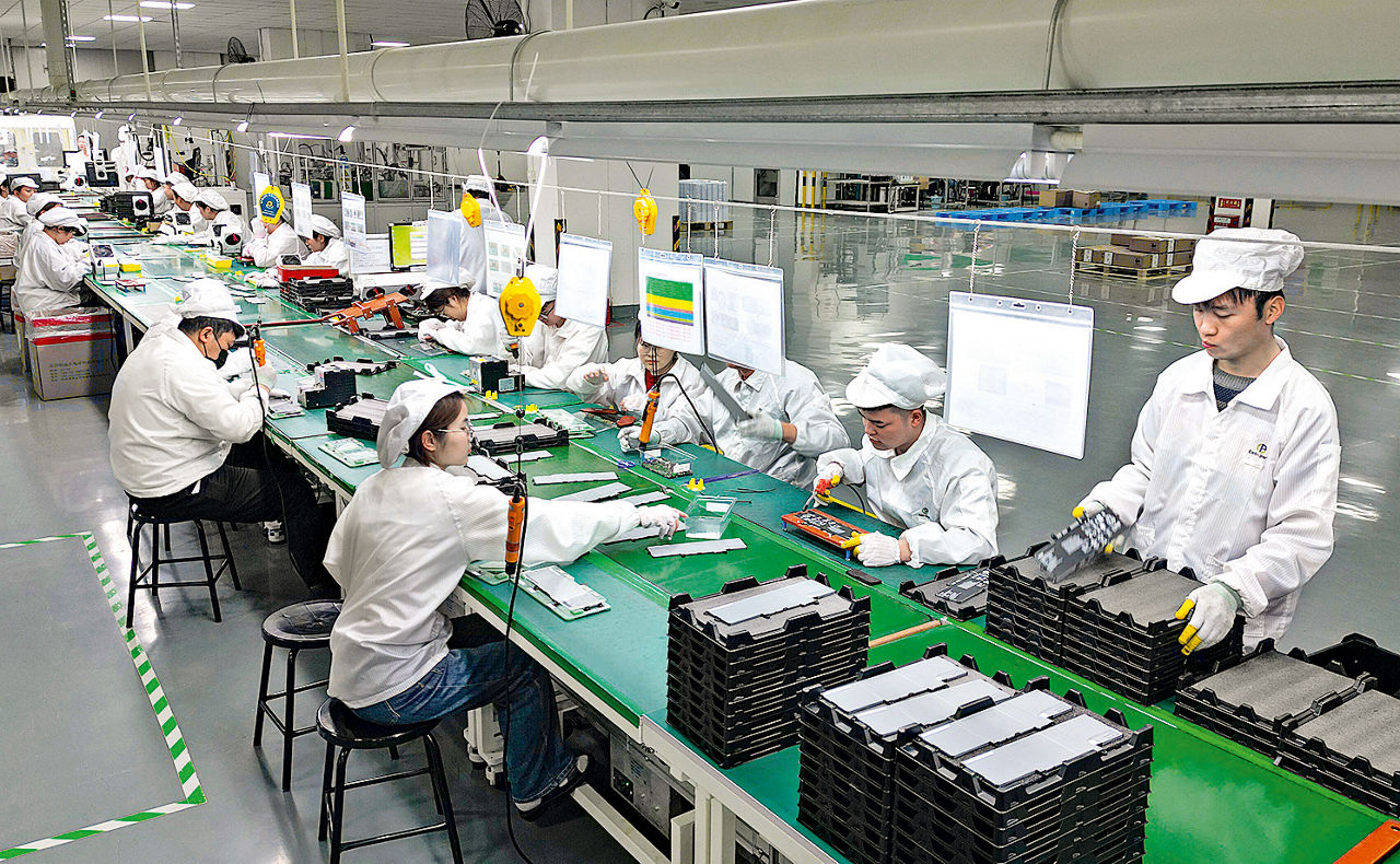 Chinese workers dressed in white assemble battery parts at a long workbench.