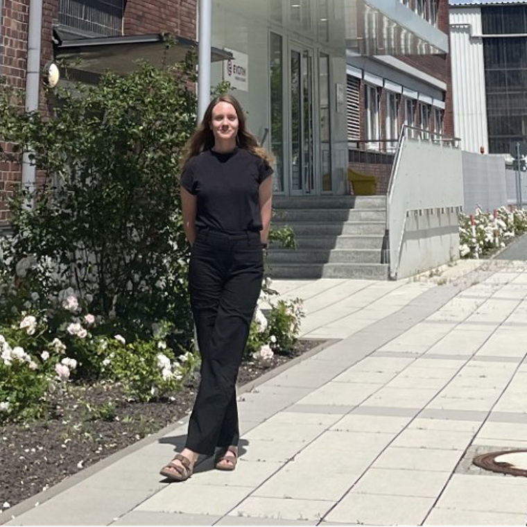 A person stands on a paved walkway beside a brick building with flags and white flowers.
