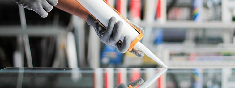 Hands wearing gray gloves applying white sealant with a caulking gun on a glass surface.