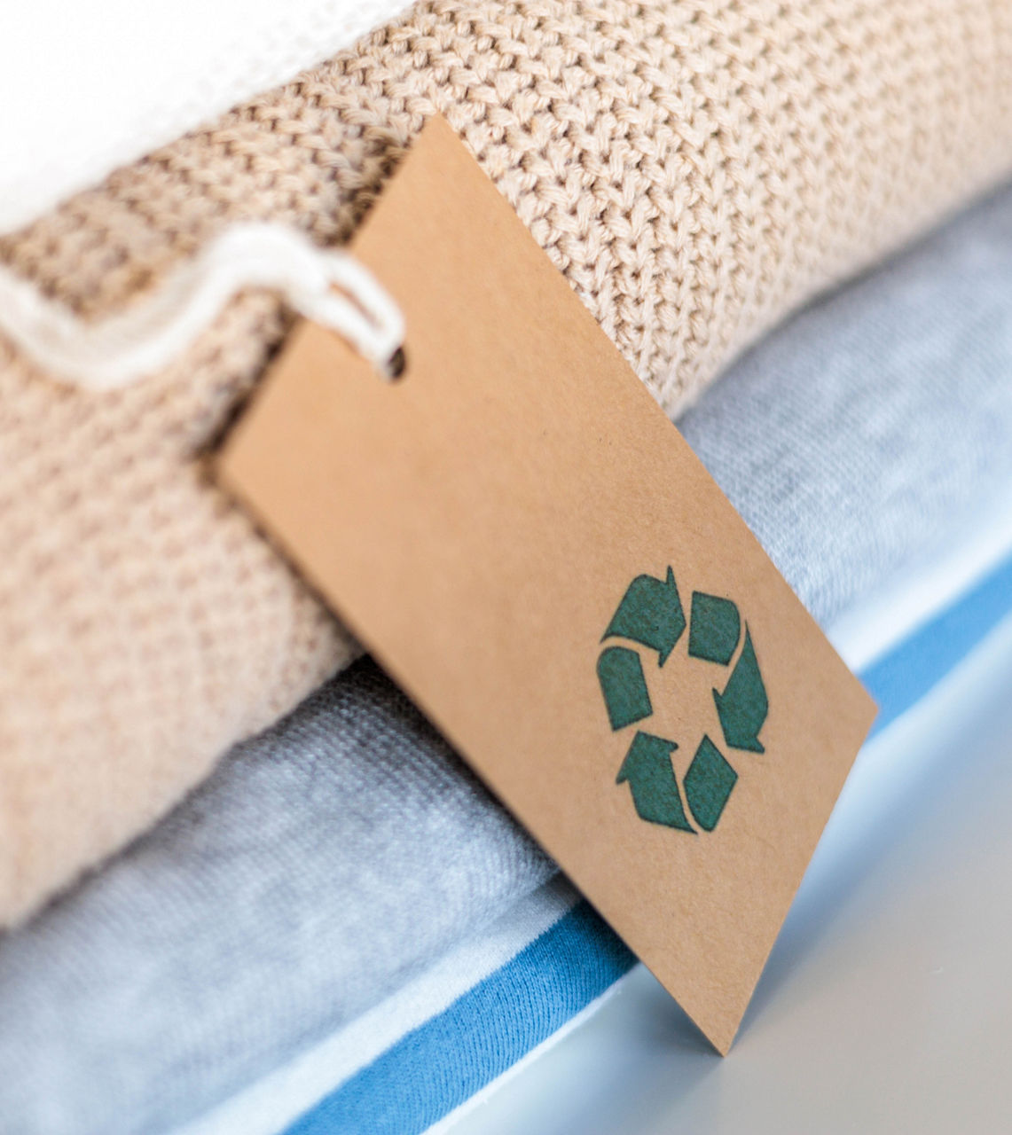 Knitted warm clothes neatly folded heap with craft tag recycling material circle arrow closeup. Eco friendly fashion clothing laundry garment made by recycled zero waste renewable textile technology