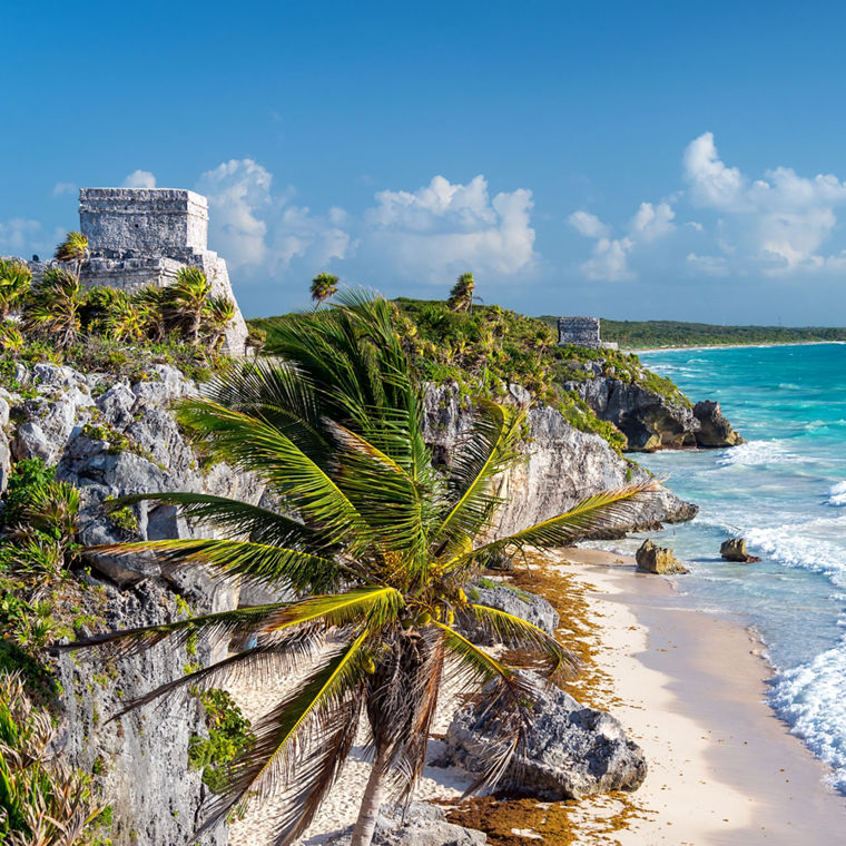 Die Ruinen von Tulum mit Blick auf das Karibische Meer an der Riviera Maya. Die Ruinen liegen erhöht auf einem Felsen, vorn eine Palme, rechts türkisblaues Meer und Sandstrand.