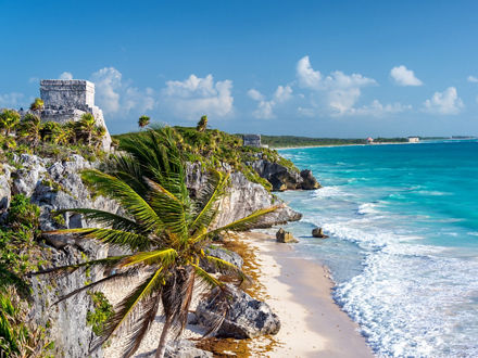 The ruins of Tulum overlooking the Caribbean Sea on the Riviera Maya (the coastal area of the Yucatán Peninsula in Mexico). The ruins are located on a cliff, with a palm tree in front, the turquoise blue sea and a sandy beach to the right.