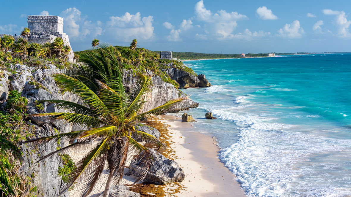The ruins of Tulum overlooking the Caribbean Sea on the Riviera Maya (the coastal area of the Yucatán Peninsula in Mexico). The ruins are located on a cliff, with a palm tree in front, the turquoise blue sea and a sandy beach to the right.