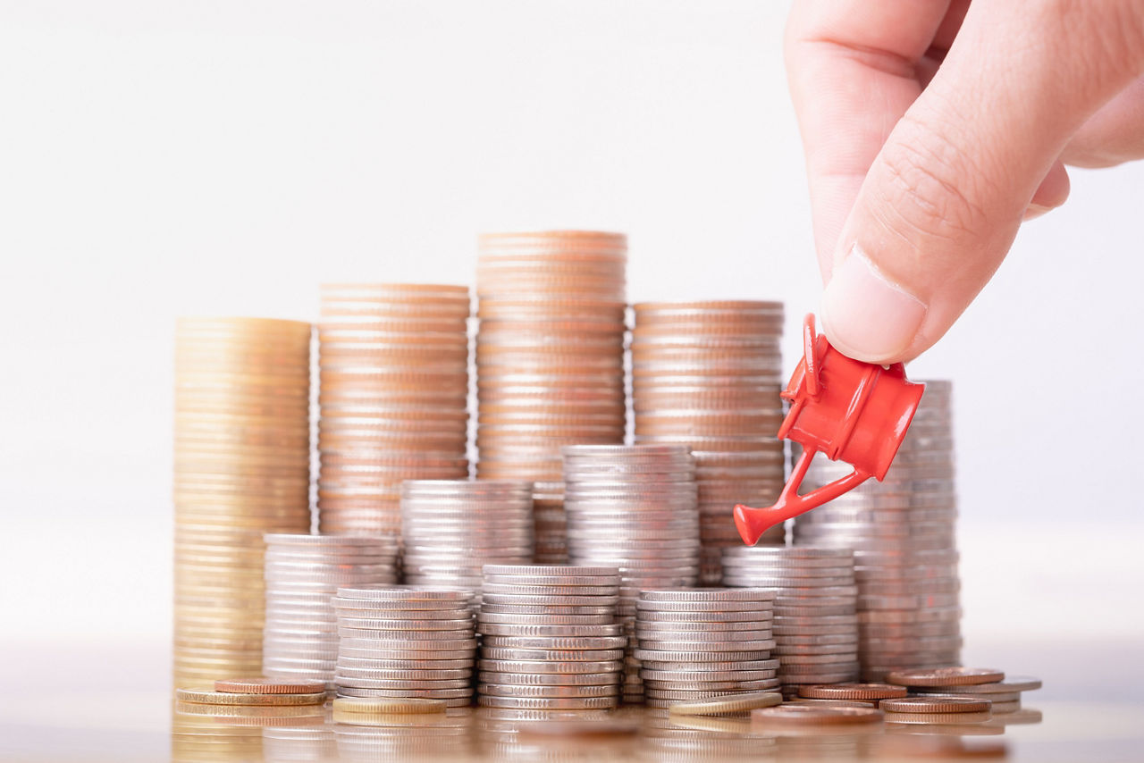 Symbolic image: Stack of coins that appear to be poured by a woman's hand using a miniature watering can.