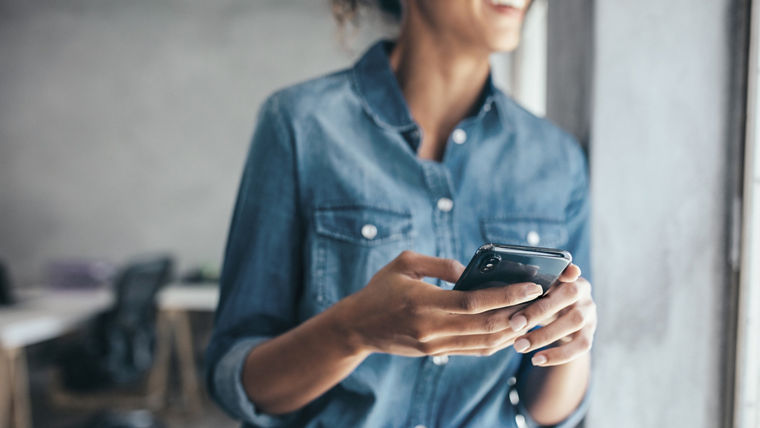 Woman in casuals standing in office by a window with phone in hand. Businesswoman with mobile phone in hand.