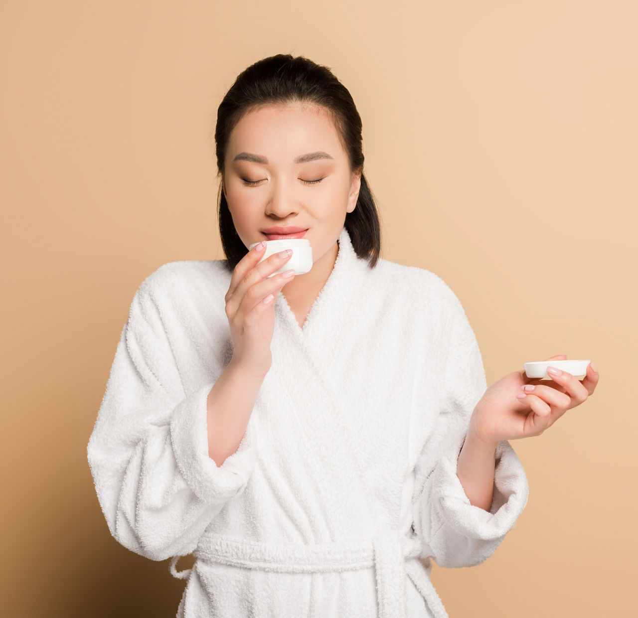 Eine lächelnde asiatisch gelesene Frau riecht an einem Creme-Tiegel. Sie trägt einen weißen Bademantel.
