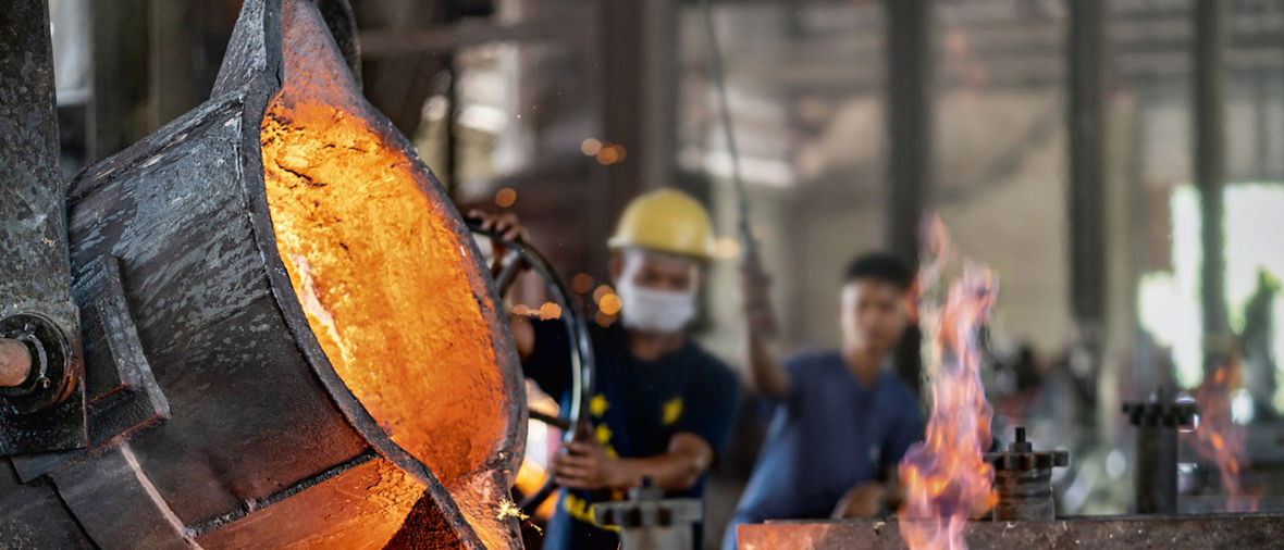 Molten metal pours from industrial ladle, worker in background.