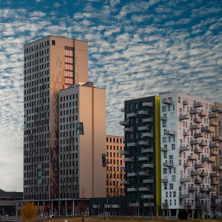 Zwei Hochhäuser nebeneinander. Das linke ist höher, hat einen etwas flacheren Anbau und hat eine Fassade aus unterschiedlich farbigen Holzflächen.