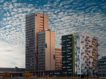 Zwei Hochhäuser nebeneinander. Das linke ist höher, hat einen etwas flacheren Anbau und hat eine Fassade aus unterschiedlich farbigen Holzflächen.