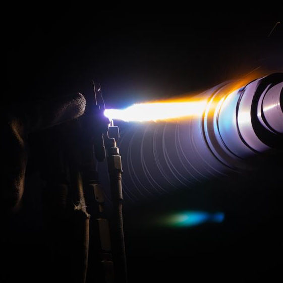 Bright flame emitting from a metal tool held by a gloved hand in a dark setting.