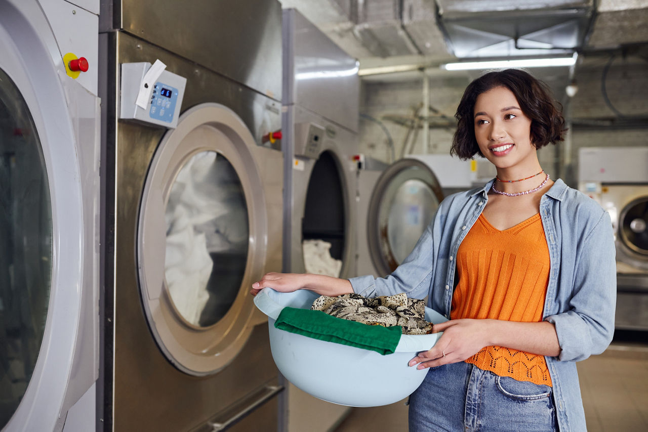 A woman with a plastic bowl full of laundry stands next to a washing machine in a laundromat.