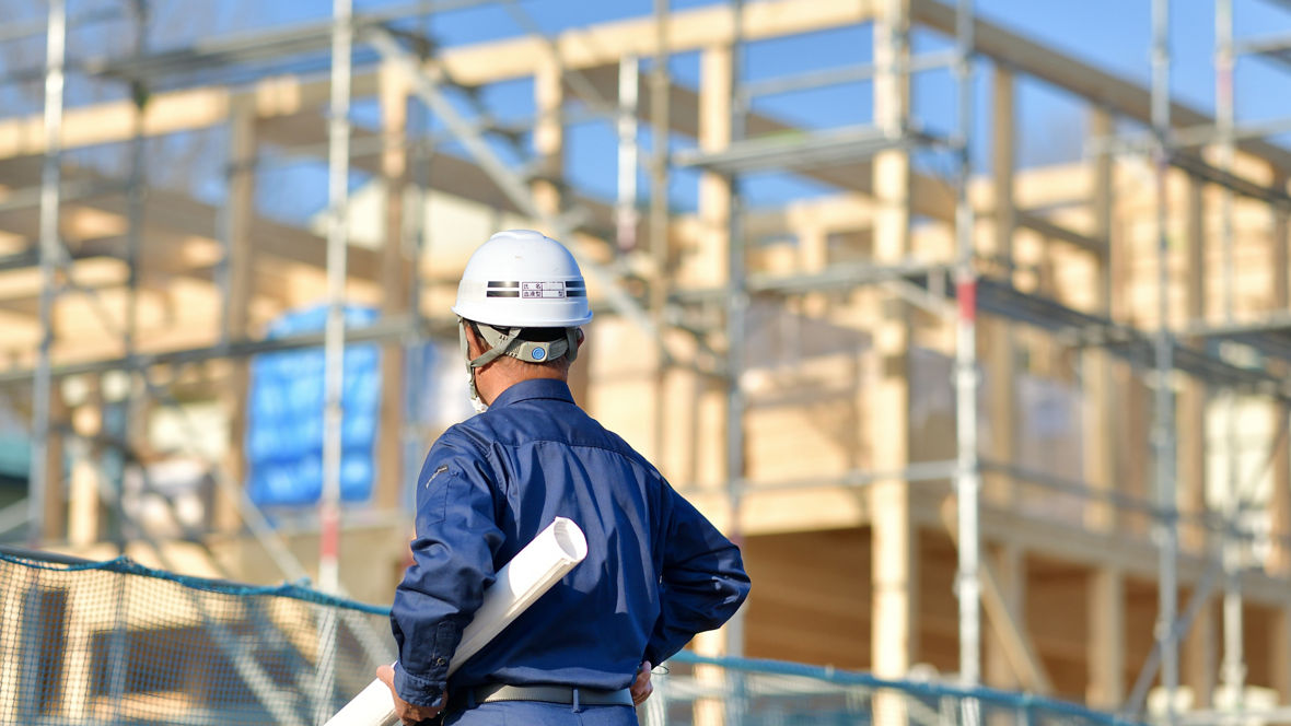 Ein Mann mit Schutzhelm blickt auf eine Holzkonstuktion für einen Neubau. Unter dem Arm trägt er einen eingerollten Bauplan.
