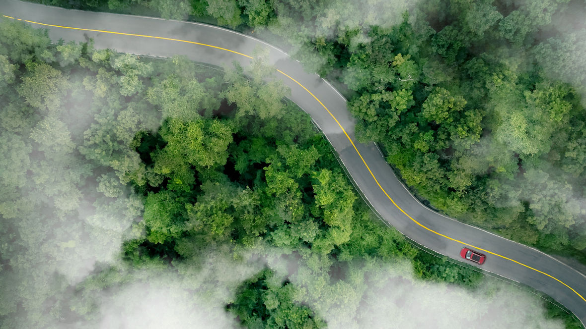 Aerial top view of a red car driving on highway road in green forest. Sustainable transport. Electric vehicle driving on asphalt road through lush green forest. Zero emission car. Green mobility.