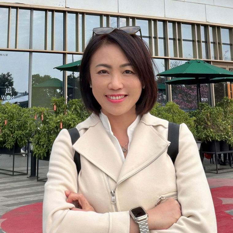 A smiling woman in a white jacket stands with arms crossed outside a modern building with green umbrellas.