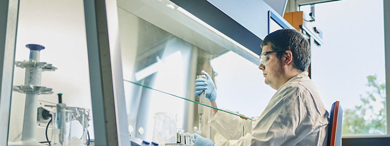 A laboratory technician wearing a white coat and safety goggles works with a pipette under a fume hood in a modern laboratory.