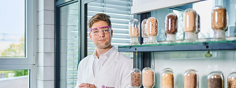 A scientist wearing a white lab coat and safety glasses holds a jar of animal feed samples. More samples are displayed on a shelf in a modern laboratory