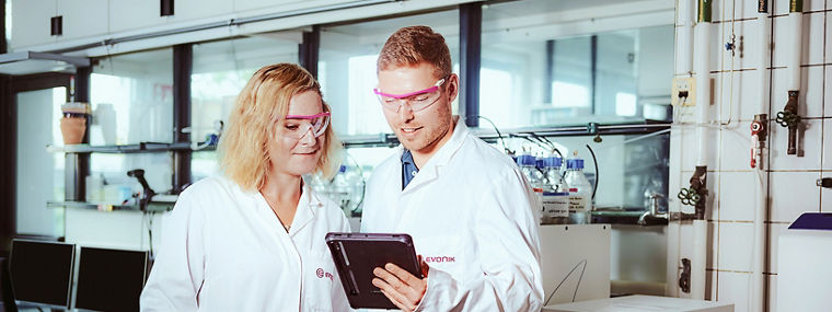 Two scientists in white lab coats and protective glasses reviewing data together on a tablet in a modern lab, discussing sustainability in animal nutrition.