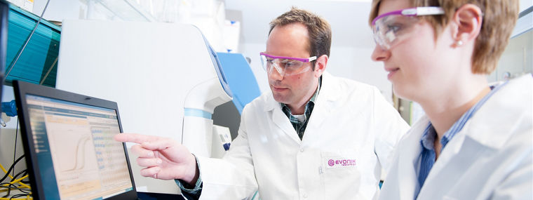 Two people wearing lab coats and safety glasses analyzing data on a screen related to animal nutrition.