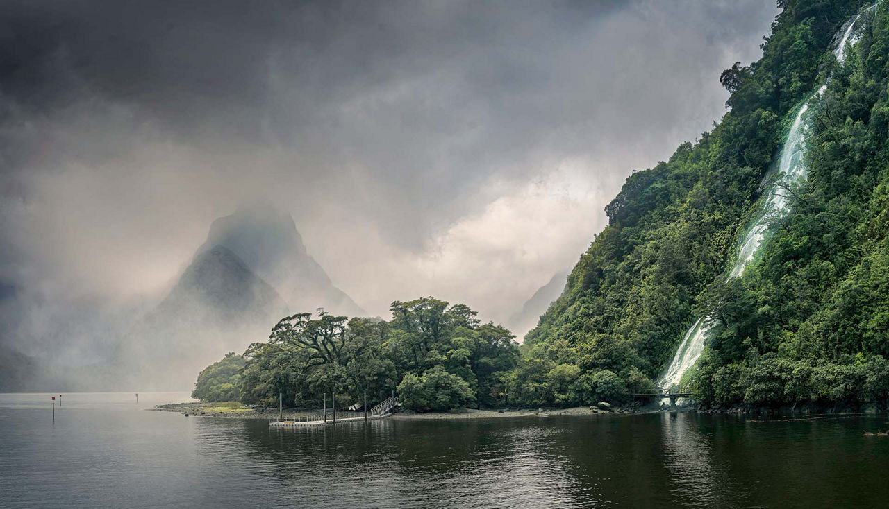 A waterfall in Milford Sound. The view extends across the water to dense clouds in the background.