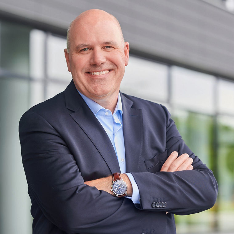 A man in a suit stands confidently with arms crossed, showcasing a professional demeanor against a modern building backdrop.