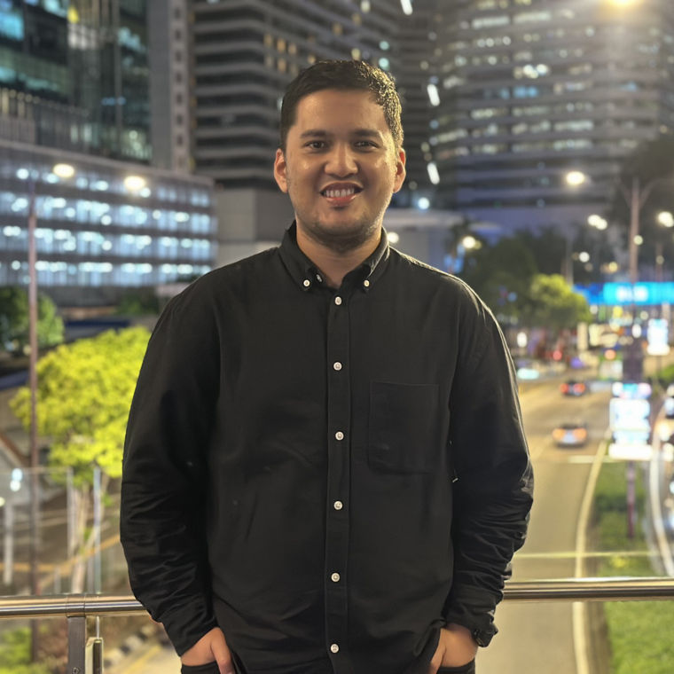 Man in a black buttoned shirt standing on a city bridge at night with blurred traffic lights.