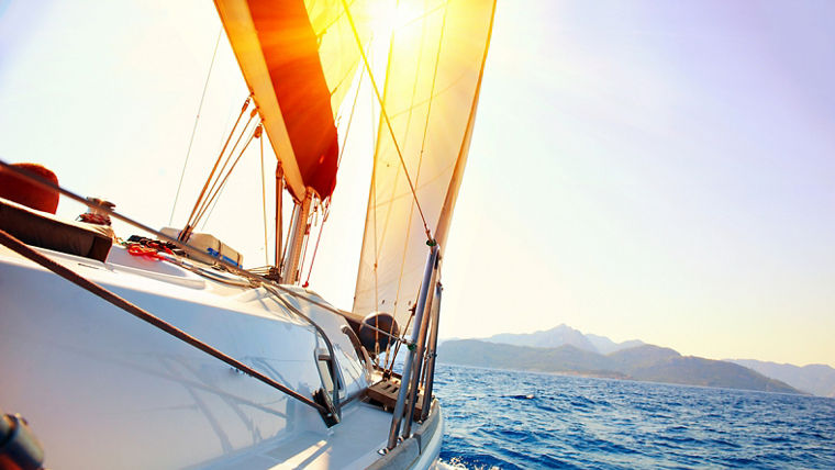 Sailboat gliding on blue ocean waters with sun shining brightly over distant mountains.