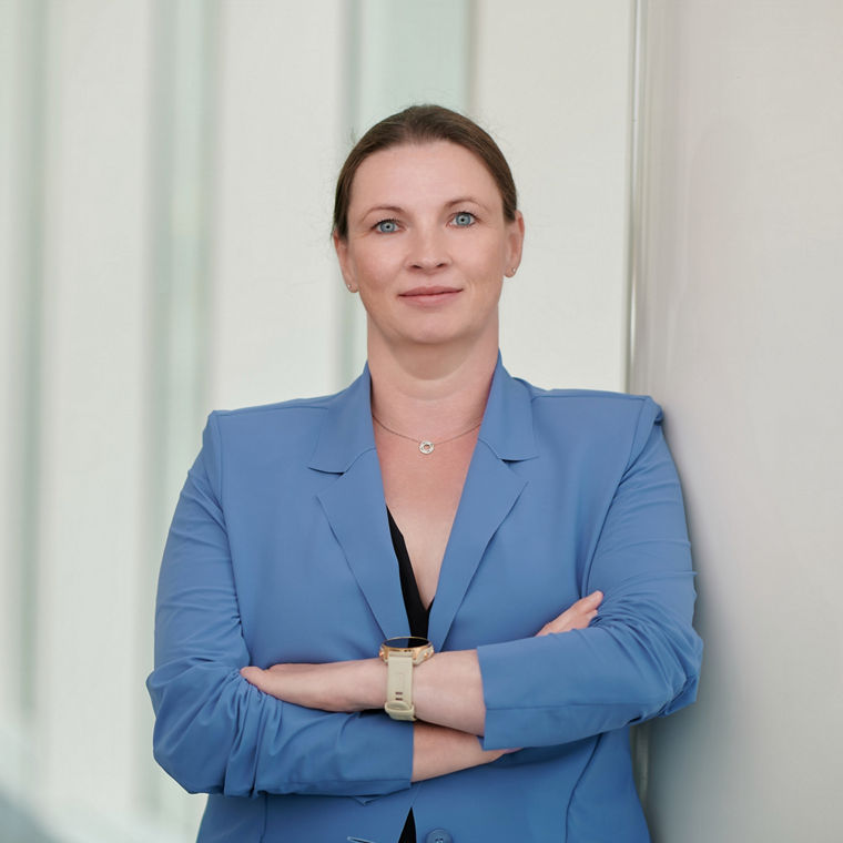 Woman with blue blazer and watch standing confidently with arms crossed against a light wall.