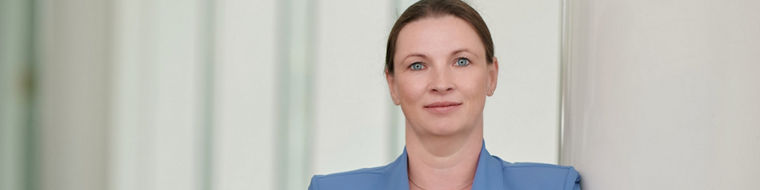 Frau mit blauem Blazer lehnt entspannt an weißer Wand mit verschränkten Armen.