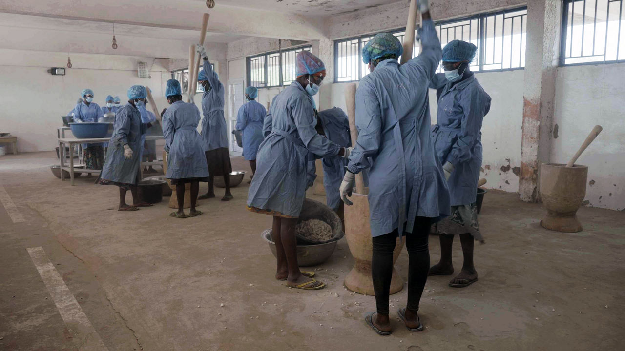 Three women in blue work coats stand around several wooden vessels, crushing the fruit pulp with wooden poles.