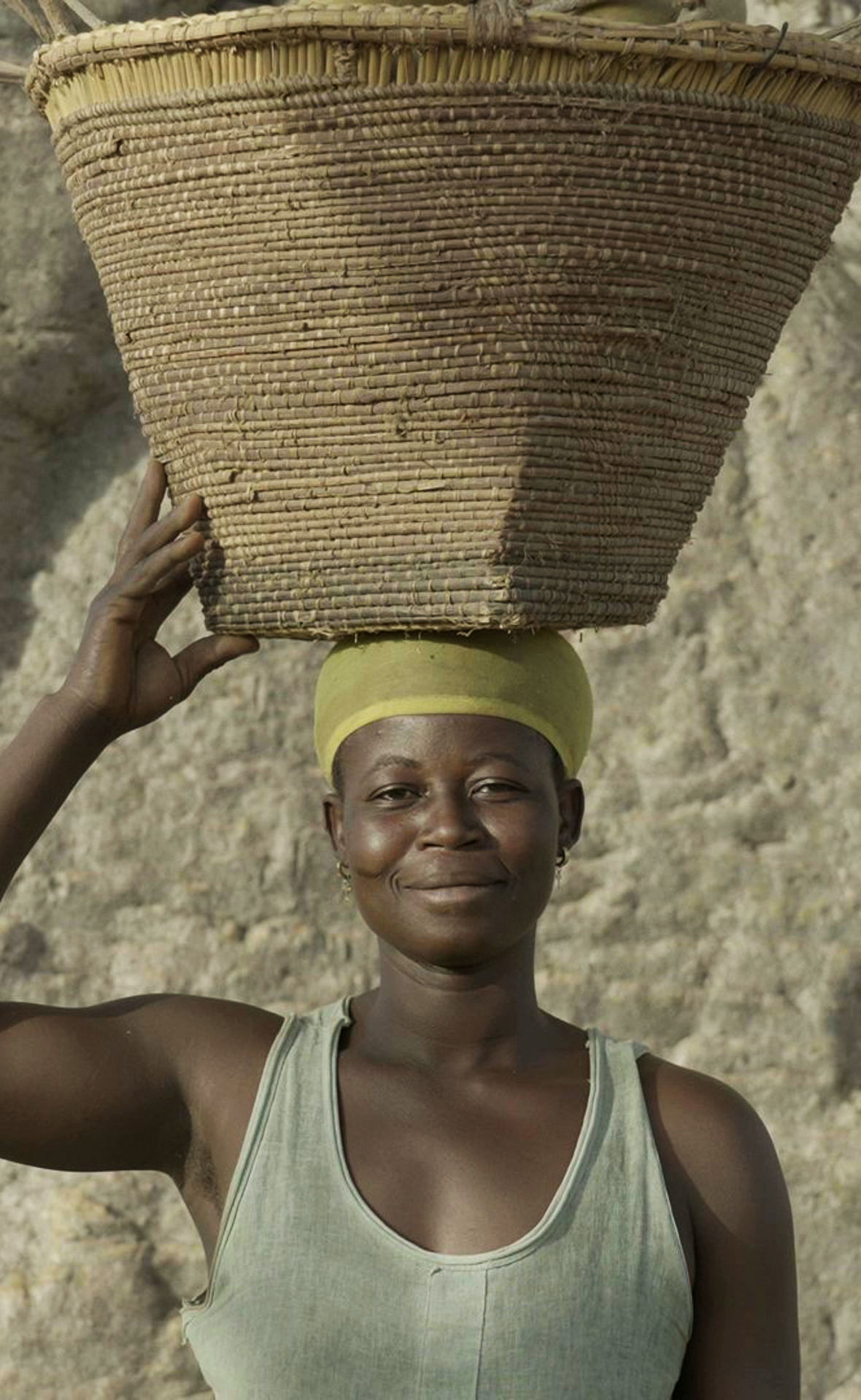 Eine Ghanaerin steht vor einem breiten Stamm eines Affenbrotbaumes. Sie trägt einen Transportkorb auf dem Kopf und lächelt in die kamera.