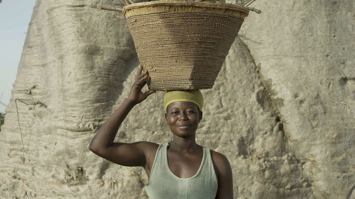 A Ghanaian woman stands in front of a wide trunk of a baobab tree. She is carrying a basket on her head and smiling at the camera.