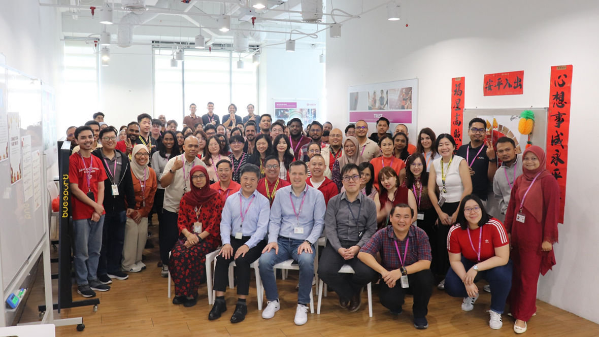 Large group of diverse colleagues posing together in a bright office space with red decorations on the wall.