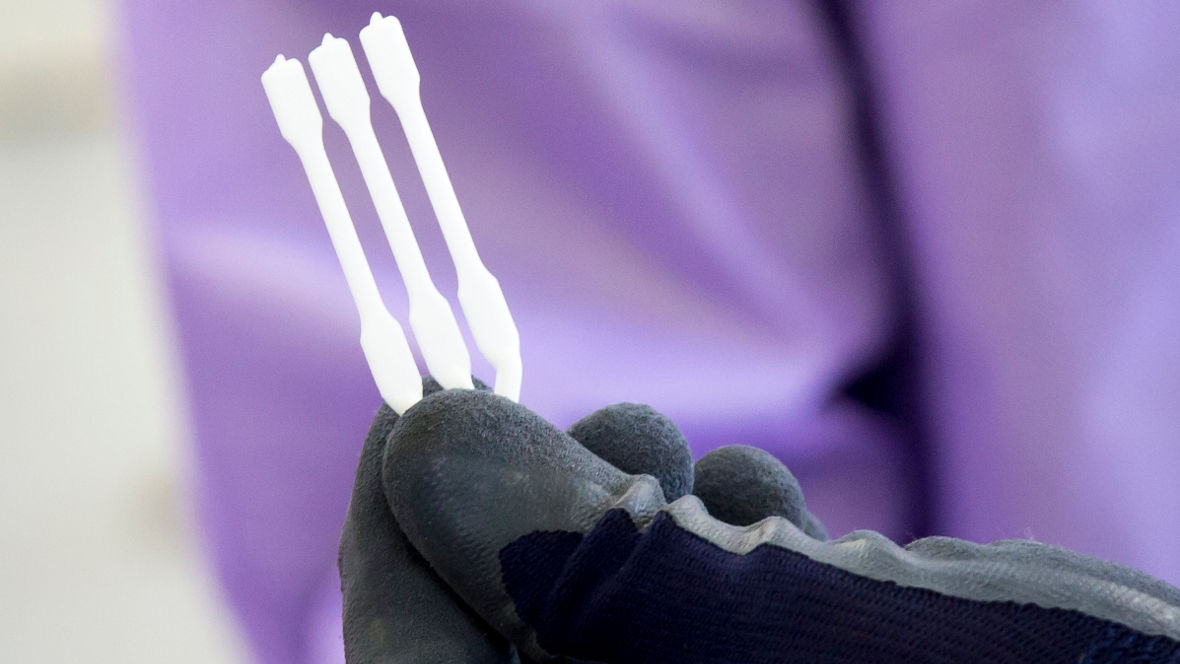 The image shows a hand wearing a black glove holding three white swabs. The swabs are thin and appear to be designed for sampling or cleaning purposes. The background is slightly blurred, with a hint of purple, which may indicate a sterile or clinical environment. The focus is on the swabs, suggesting their importance in the context of the image, possibly related to medical or laboratory procedure