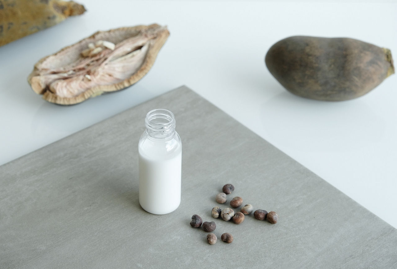 A white lotion in a bottle is standing on a table. Around it are the fruits of the baobab tree and some of its seeds.