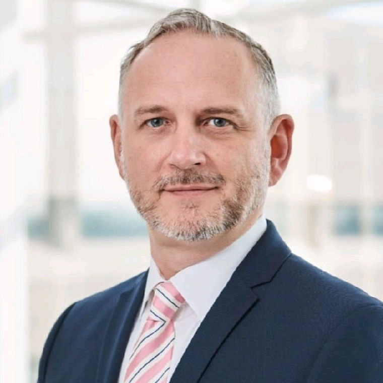 A professional man in a suit with a pink striped tie, standing in a bright, modern office environment.