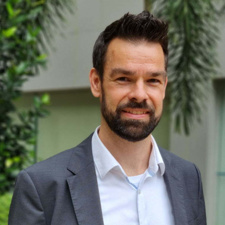 A man in a gray suit stands outdoors, surrounded by greenery, conveying a professional appearance.