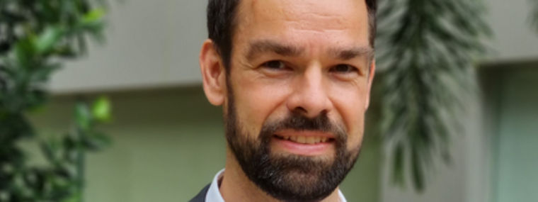 A man in a gray suit stands outdoors, surrounded by greenery, conveying a professional appearance.