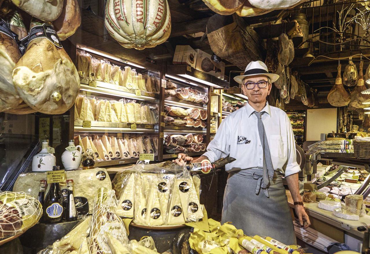 Ein Mann mit Hut zwischen Käseblöcken und von der Decke hängenden Schinken.