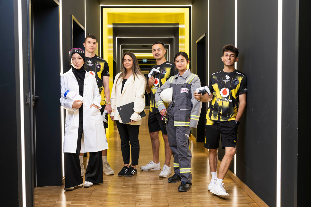 Sechs Personen in verschiedenen Berufs- und Sportkleidungen stehen in einem beleuchteten Gang.