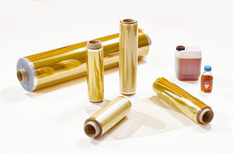Rolls of shiny gold-colored film with two containers of liquid in the background on a white surface.