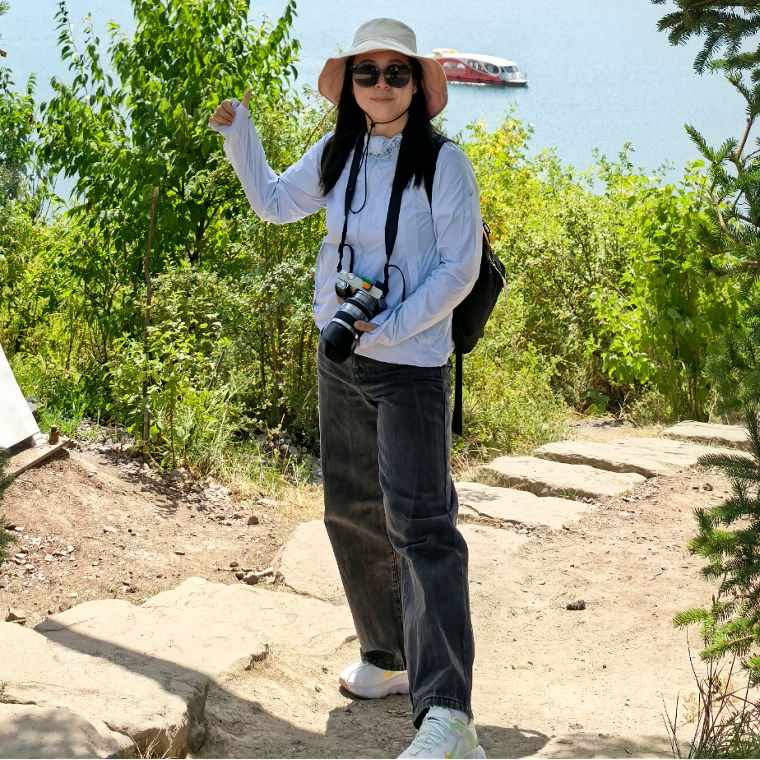 Eine Person posiert mit einer Kamera vor einem malerischen Bergsee, umrahmt von Bäumen und einer modernen Struktur.